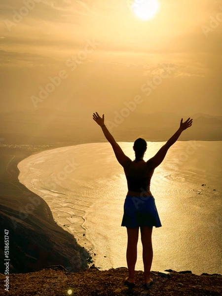 Fototapeta SILHOUETTE, LENS FLARE: Woman with raised arms overlooks a sunlit bay, with shimmering water reflecting soft glow of sky. Dramatic coastal landscape behind her evokes a sense of peace and achievement