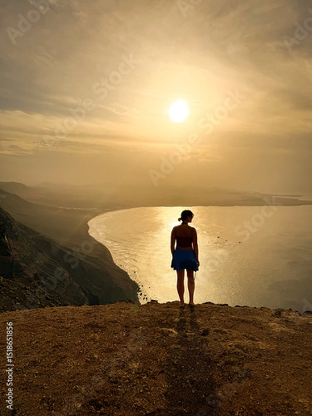Fototapeta SILHOUETTE, LENS FLARE: A lone female figure stands at a vantage point, observing a vast coastal bay framed by distant mountains. Stunning view from a clifftop of Famara beach in soft morning light.