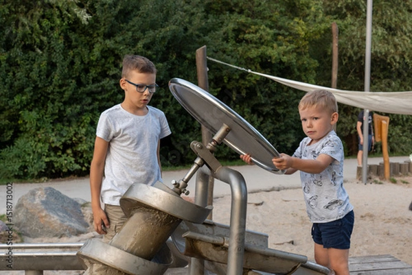Obraz Focused young boys playing with interactive water screw pump at outdoor playground on sunny day. Concept of childhood curiosity, teamwork, educational play, and public park activities.