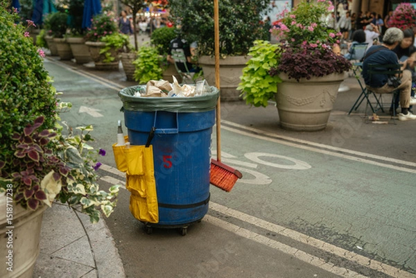 Obraz A blue trash bin with a broom and cleaning supplies stands on a street in New York. Large planters and people are seen in the background of this urban setting.