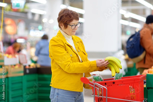 Fototapeta Beautiful red-haired senior lady is choosing fresh organic bananas in a food store or a supermarket .