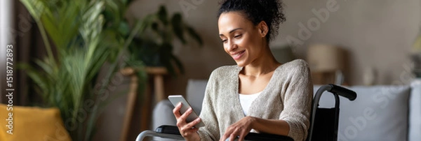 Fototapeta Happy woman in wheelchair is sitting indoors and smiling while using smartphone, enjoying relaxed moment at home
