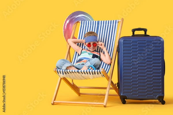 Fototapeta Cute little boy with inflatable ring and suitcase sitting in deck chair on yellow background. Travel concept
