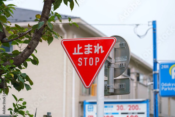 Fototapeta Tokyo, Japan - Sep 30 2024, A close-up view of a triangular red traffic stop sign hanging over the road, with building facades and an cloudy sky in the background, Tokyo, Japan