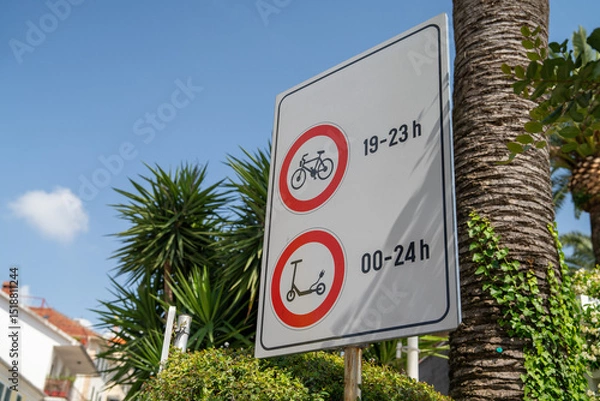 Fototapeta Sign showing time restrictions for bicycles and scooters, set against a vibrant tropical backdrop, featuring lush palm trees and clear blue sky.