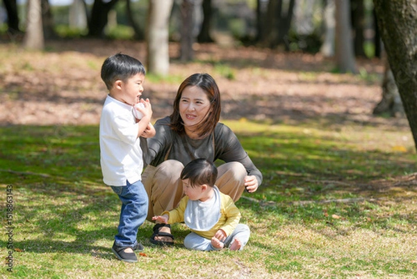 Fototapeta In March, at a sunny forested park in Japan, a Japanese mom and her 6-month-old baby sit on the grass. Nearby, her 3-year-old son tosses a ball. This photo captures a peaceful spring family moment.