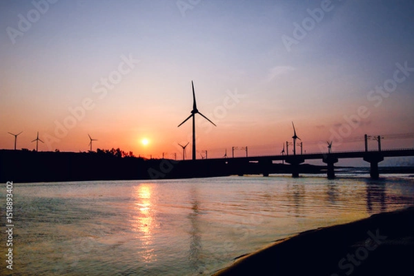 Obraz Silhouetted windmills and a bridge are visible against a vibrant sunset over the water in Houlong Port, Miaoli County, Taiwan. Capturing the beauty of renewable energy in coastal landscapes.