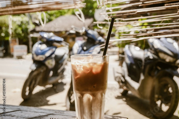 Fototapeta Refreshing iced coffee latte served in a glass with straw outdoor at daytime on the terrace of a restaurant in General Luna on Siargao in the Philippines with blurry background.