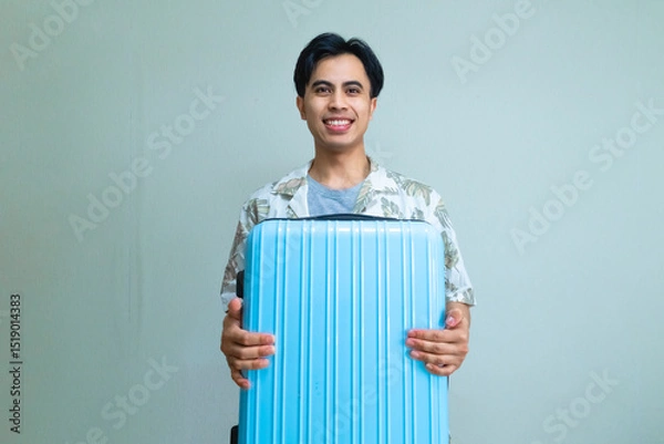 Fototapeta Smiling young Asian man in summer clothes carrying and hugging a suitcase, looking cheerful and ready for vacation, posing against a clean, colorful background for travel or holiday concepts