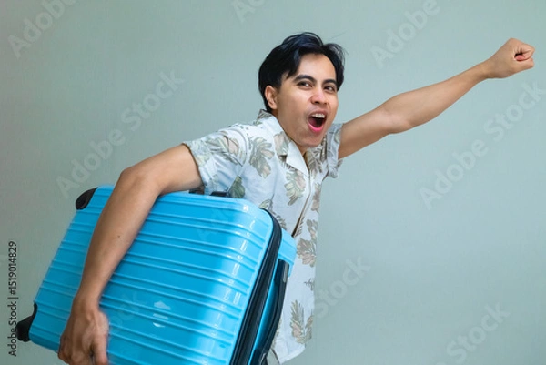 Fototapeta Happy young Asian man in summer shirt shouting "GO" while carrying a bright blue suitcase, posing with excitement against a plain background, ready for a fun holiday or travel adventure