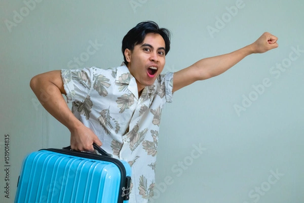 Fototapeta Happy young Asian man in summer shirt shouting "GO" while carrying a bright blue suitcase, posing with excitement against a plain background, ready for a fun holiday or travel adventure