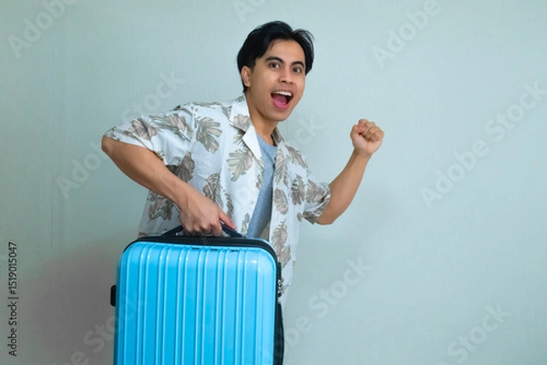 Fototapeta Excited young Asian man in summer clothes posing with a running gesture while holding a blue suitcase, isolated on a clear background, ready for vacation or travel adventure