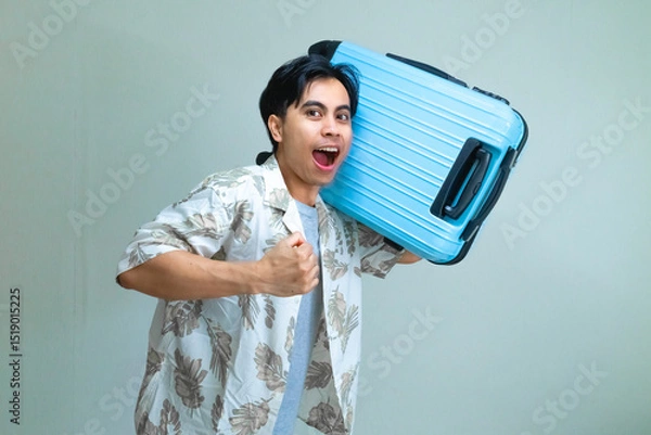Fototapeta Excited young Asian man in summer clothes posing with a running gesture while holding a blue suitcase, isolated on a clear background, ready for vacation or travel adventure