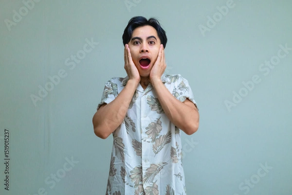Fototapeta Portrait of a young Asian man in summer clothes with an amazed expression, posing with a surprised hand gesture against a clear background, perfect for promotion or advertisement