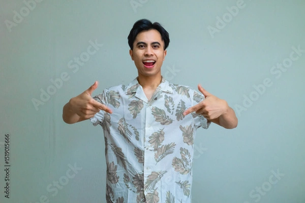 Fototapeta Happy young Asian man in summer outfit smiling and pointing down with both hands, standing against a clear background, perfect for promotions, ads, or social media content