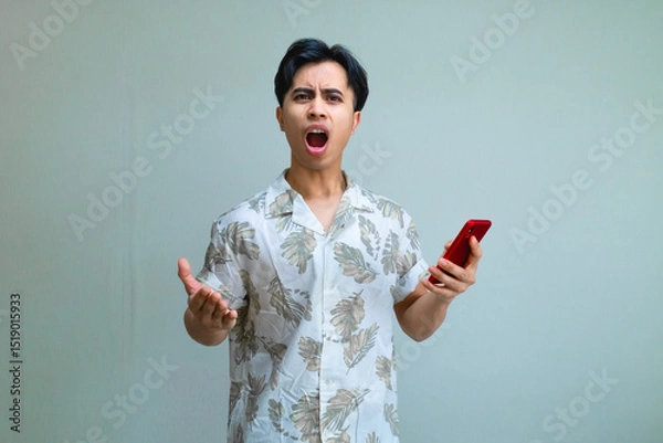 Fototapeta Angry young Asian man in summer clothes holding a smartphone and staring at the camera with a serious expression, isolated on a clean studio background