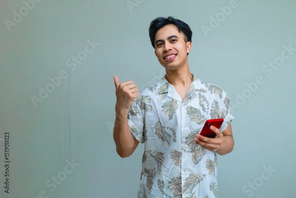 Fototapeta Happy young Asian man in summer clothes pointing aside with his thumb while holding a smartphone, isolated on a clear background, showing excitement and joy