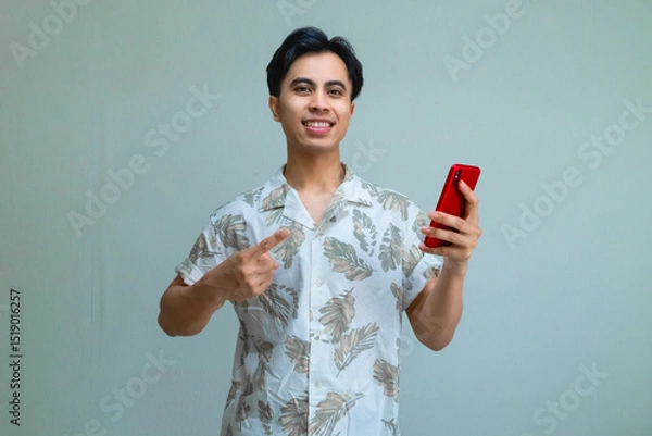Fototapeta Portrait of a smiling young Asian man in summer clothes pointing at a cellphone he is holding, isolated on a clear background, perfect for digital ads, promotions, or mobile app concepts