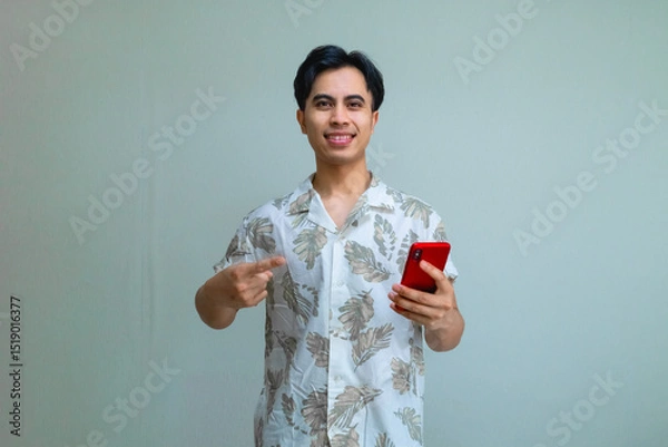 Fototapeta Portrait of a smiling young Asian man in summer clothes pointing at a cellphone he is holding, isolated on a clear background, perfect for digital ads, promotions, or mobile app concepts