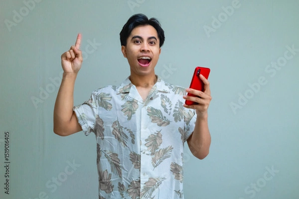 Fototapeta Excited young Asian man in summer clothes pointing up while holding a cellphone, standing against a clear background, showing cheerful mood and vibrant holiday energy