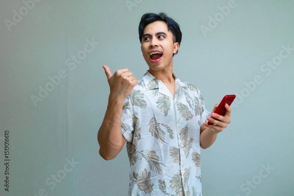 Fototapeta Happy young Asian man in summer clothes pointing aside with his thumb while holding a smartphone, isolated on a clear background, showing excitement and joy