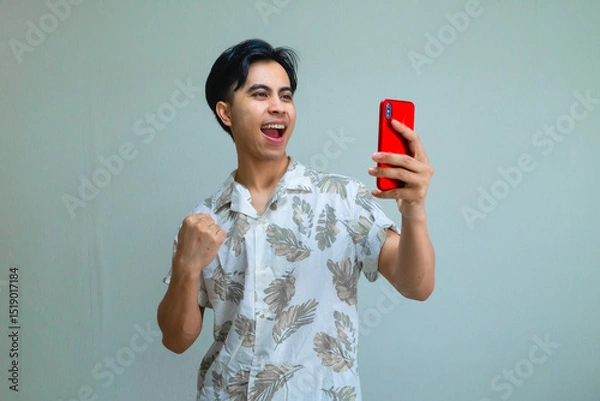 Fototapeta Happy young Asian man wearing summer clothes celebrates success with clenched fist while holding smartphone, excited and smiling on a clear background, ideal for promotions