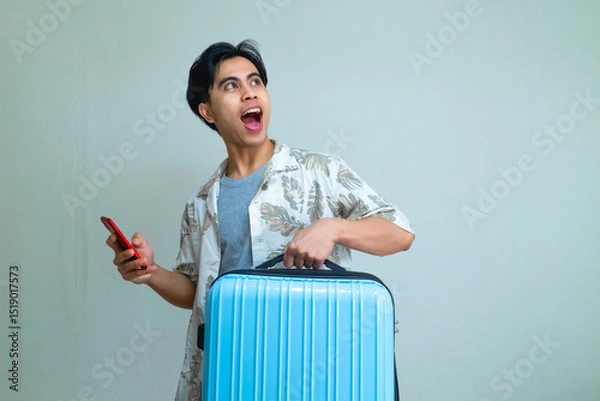 Fototapeta Happy young Asian man in summer clothes looking to the side with an excited expression, holding a smartphone and suitcase, standing against a clear background with copy space