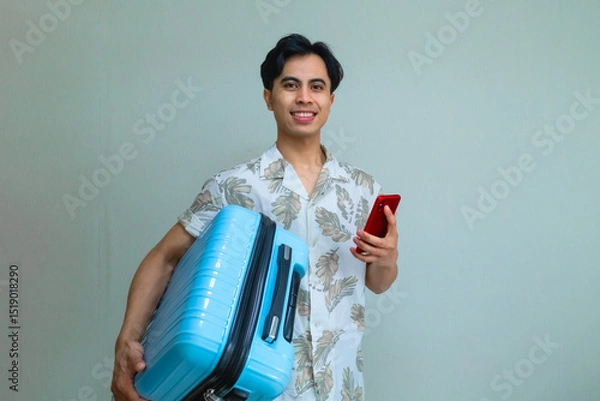 Fototapeta Happy young Asian man in summer outfit smiling while using smartphone and carrying a blue suitcase, ready for travel or vacation, isolated on a clear background