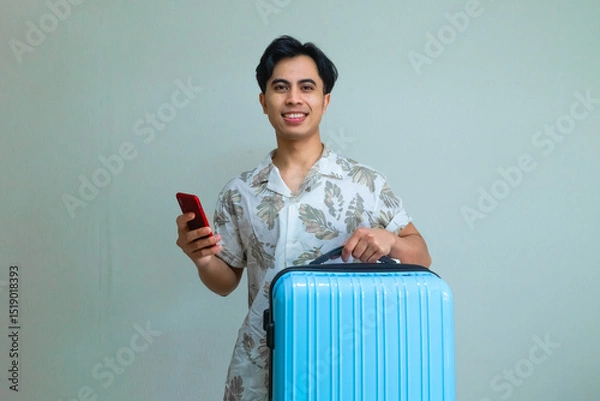 Fototapeta Happy young Asian man in summer outfit smiling while using smartphone and carrying a blue suitcase, ready for travel or vacation, isolated on a clear background