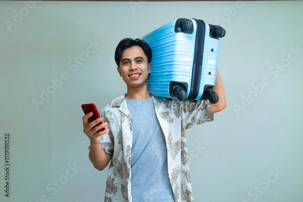 Fototapeta Happy young Asian man in summer outfit smiling while using smartphone and carrying a blue suitcase, ready for travel or vacation, isolated on a clear background