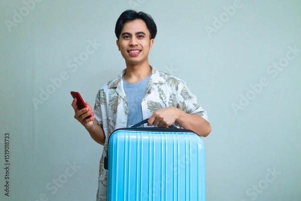 Fototapeta Happy young Asian man in summer outfit smiling while using smartphone and carrying a blue suitcase, ready for travel or vacation, isolated on a clear background