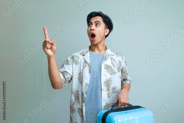 Fototapeta Portrait of a young Asian man in summer clothes pointing to the side with a funny wow expression while carrying a suitcase, isolated on a clear background, travel concept