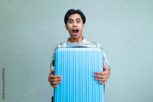Fototapeta Happy young Asian man in summer clothes hugging a suitcase with a wow expression, looking at the camera, isolated on a clear background, ready for vacation or travel