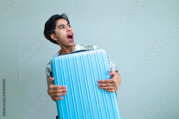 Fototapeta Portrait of a young Asian man in summer outfit looking to the side with a surprised wow expression while hugging a suitcase, isolated on a clear background, travel concept