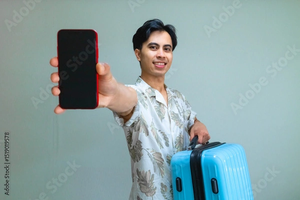 Fototapeta Smiling young Asian man in summer outfit showing blank mobile phone screen and carrying a blue suitcase, perfect for travel app, booking service, or holiday advertisement concept