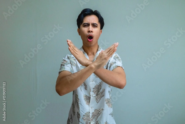 Fototapeta Portrait of a serious young Asian man in summer clothes making a stop gesture with his hand, isolated on a clear background, expressing warning or refusal