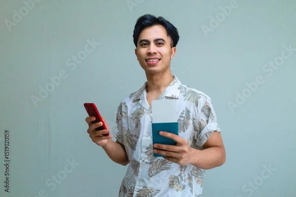 Fototapeta Happy young Asian man in summer outfit holding passport and plane ticket while using smartphone, excited for vacation or travel, isolated on a clear background