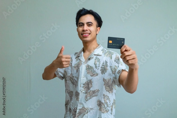 Fototapeta Smiling young Asian man wearing summer clothes holds a product card and gives a thumbs up, perfect for advertisement on a clear background