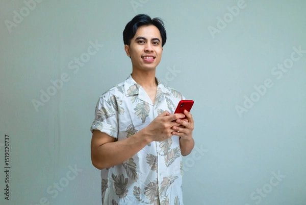 Fototapeta Portrait of a smiling young Asian man wearing summer clothes, confidently using a cell phone on a clean clear background, radiating a relaxed and positive mood