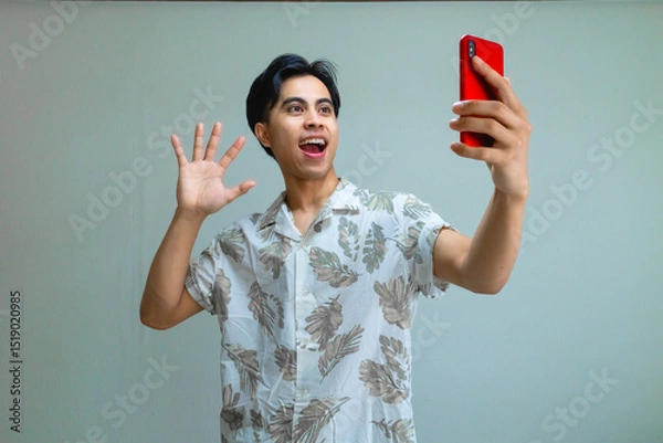 Fototapeta Happy young Asian man in summer outfit recording himself with a smartphone, smiling and making a fun gesture against a clean, clear background, perfect for social media or advertisement use