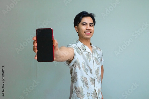 Fototapeta Happy young Asian man in summer outfit showing a blank mobile phone screen, standing against a clean background, perfect for advertising, app promo, or digital marketing