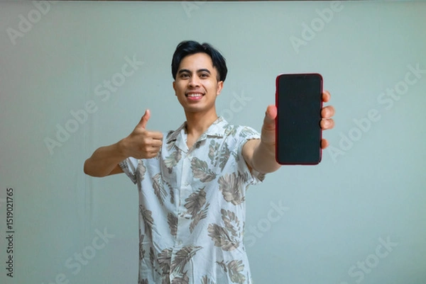 Fototapeta Happy young Asian man in summer outfit giving a thumbs-up while showing a blank mobile phone screen, standing against a clear background, perfect for app promo or advertisement