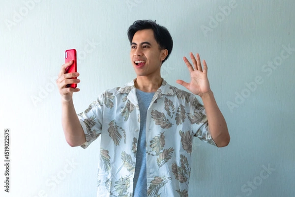 Fototapeta Portrait of a smiling young Asian man in summer clothes making a video call gesture, isolated on a clear background, cheerful and friendly expression perfect for digital ads or promotions