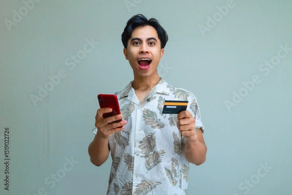 Fototapeta appy young Asian man in summer outfit holding a credit card and using a smartphone for online transaction, showing amazed face expression against a clear background