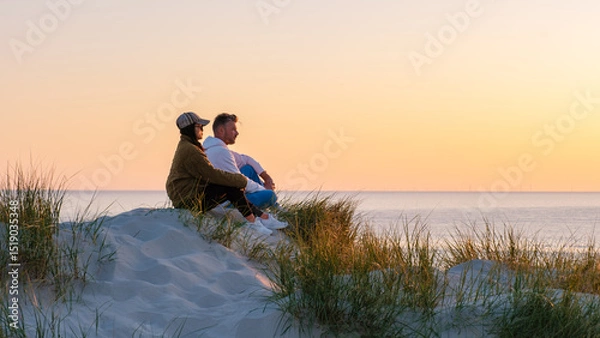 Fototapeta A couple sits on a sandy dune, surrounded by tall grass, admiring the vibrant sunset over the calm waters of the beach in Denmark. The warm hues create a romantic atmosphere. Vejers Strand beach