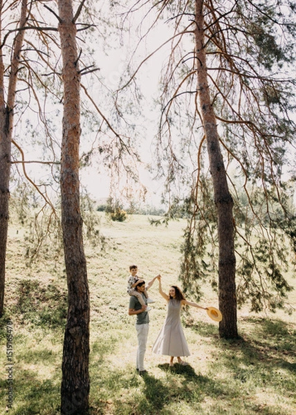 Obraz Young family walking through park, mother dancing while child rides on father’s shoulders.