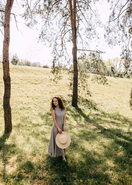 Obraz Woman in long dress holding straw hat, standing on sunny grassy hill under trees. Peaceful moment in nature with warm light and soft shadows.