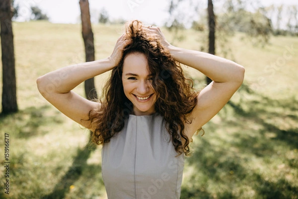 Fototapeta Joyful young woman with curly hair holding her head and smiling brightly in a sunlit park, surrounded by trees and grass.