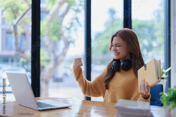 Fototapeta An excited woman celebrates success while working from home with laptop.
