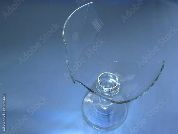Fototapeta A broken wine glass with a wedding ring inside, placed on a blue background. Conceptual image symbolizing relationship breakup, vulnerability, betrayal, or loss of love.